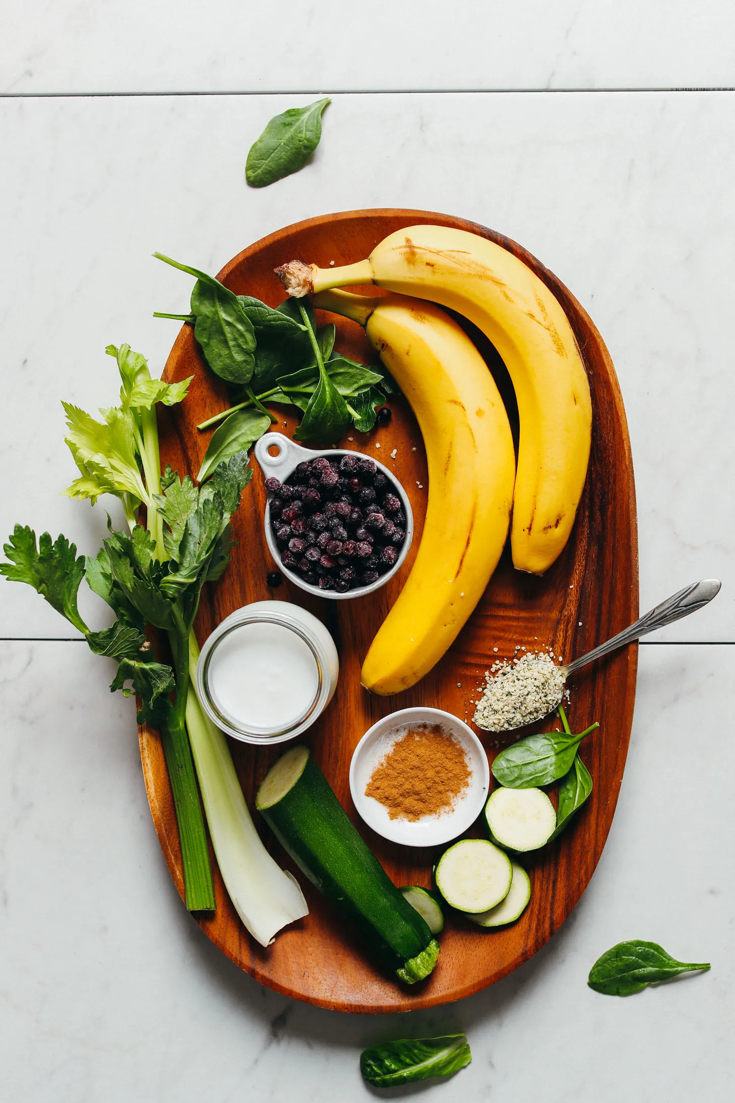 Celery, zucchini, coconut milk, blueberries, spinach, banana, hemp seeds, and cinnamon displayed on a wood platter